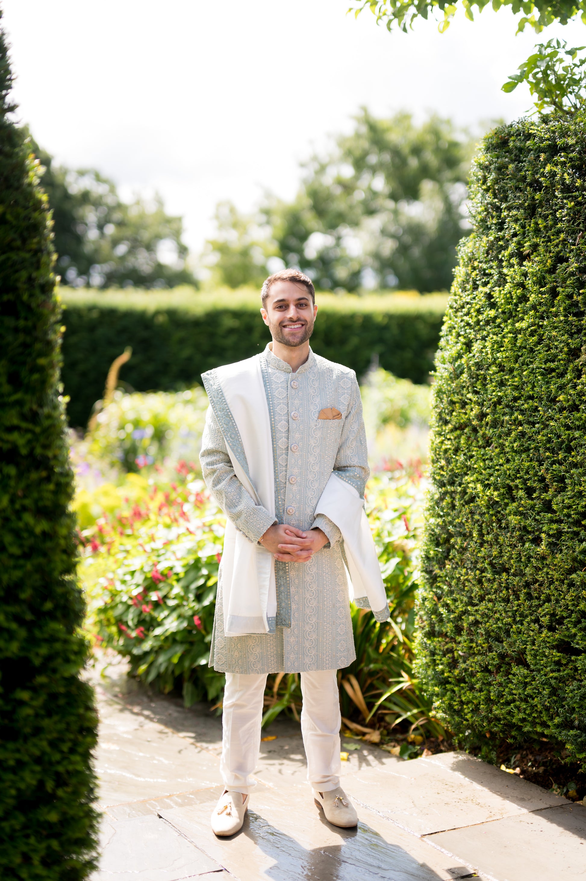 Groom in ice blue sherwani and white trousers, accessorized with off-white/cream suede Mojaris/Mojris, surrounded by trees